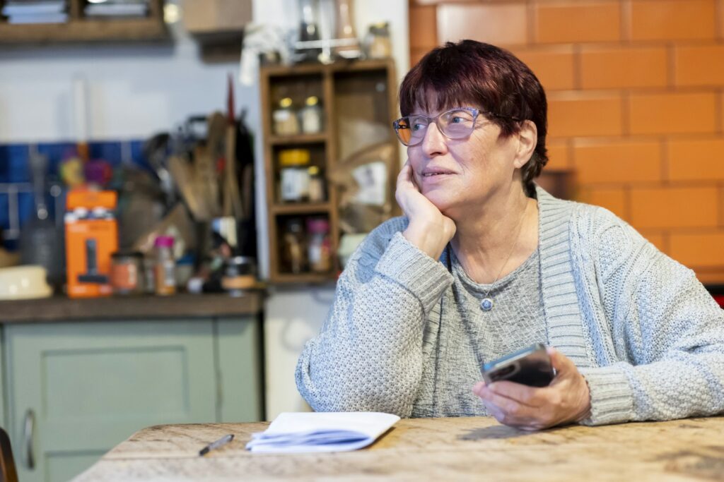 Idosa sentada à mesa com celular e papéis, preocupada
